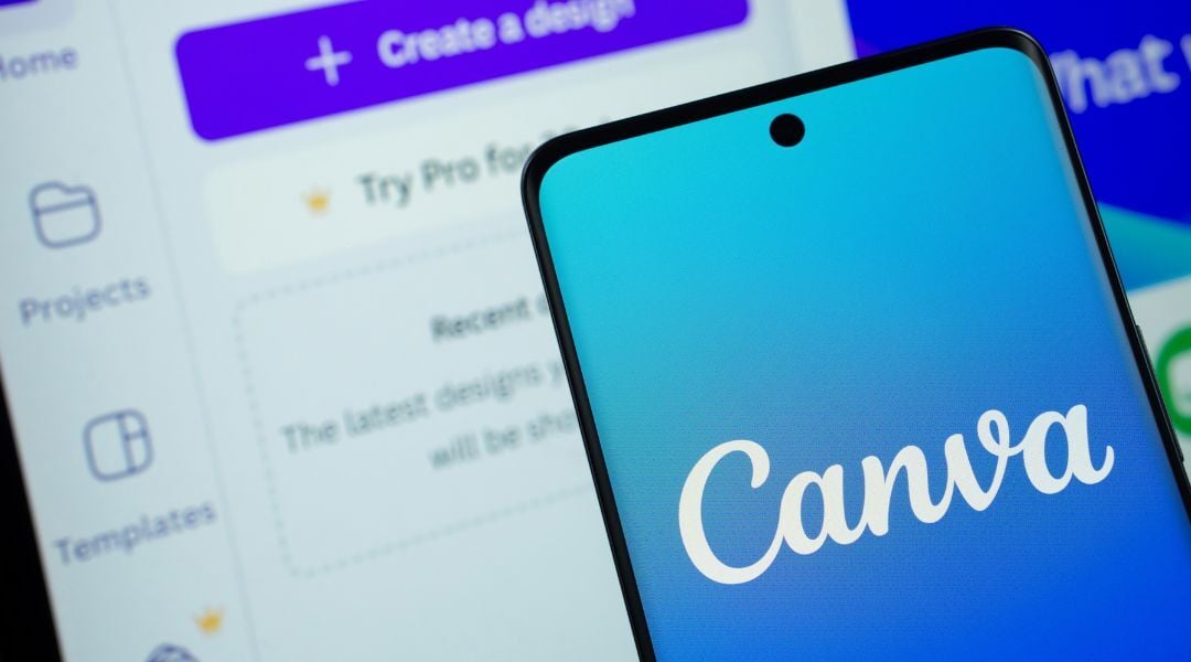 A close-up shot of a smartphone screen displaying the Canva logo, held in front of a computer monitor showing the Canva website.