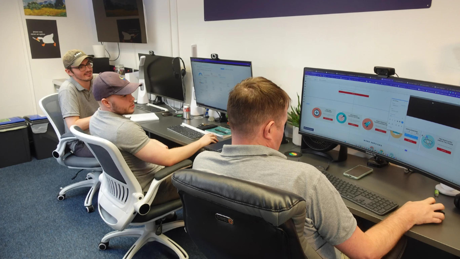 Our old office which shows Michael, Leon and Toby all sitting at their desks, which are graphite grey and each desk has black monitors, with very little space 