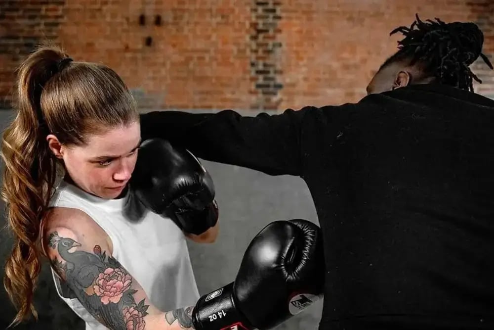 This image shows two people boxing the person in the foreground is wearing a white t-shirt and black boxing gloves while the person she is sparring with has their back turned to the camera and is wearing a black long sleeve top.