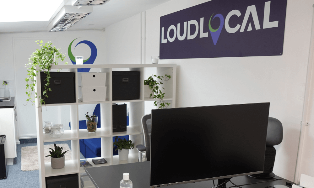 Our first office in Kenilworth, Warwickshire with the LoudLocal branding in the background.  Showing a grey desk, with a lamp and a white shelving unit