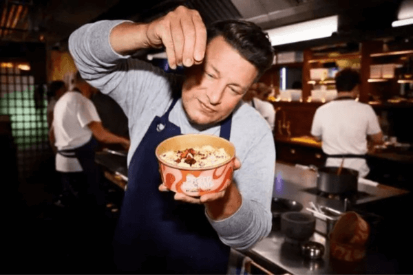 An image of Jamie Oliver who is the owner of the brand Pasta Dreams, wearing a grey jumper and blue apron sprinkling cheese on top of a pasta dish in a takeaway box.