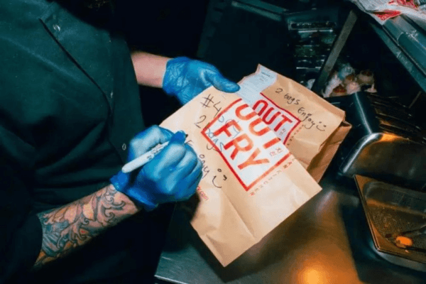 Taster is one of LoudLocal's clients. This image shows a person wearing blue kitchen gloves writing on brown paper bags with Out Fry written on the outside