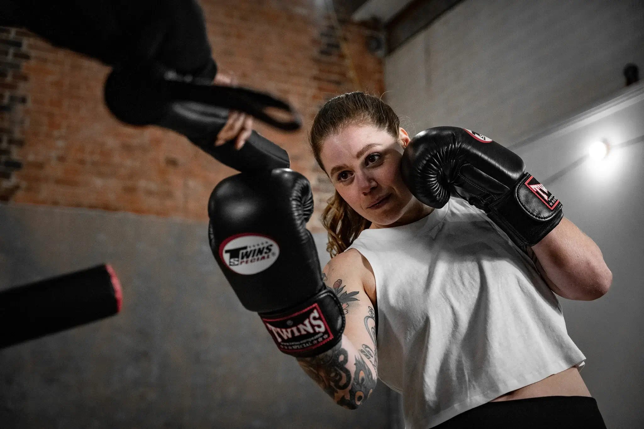 This image shows a woman wearing a white t-shirt and black boxing gloves training with a coach at Bronx gym