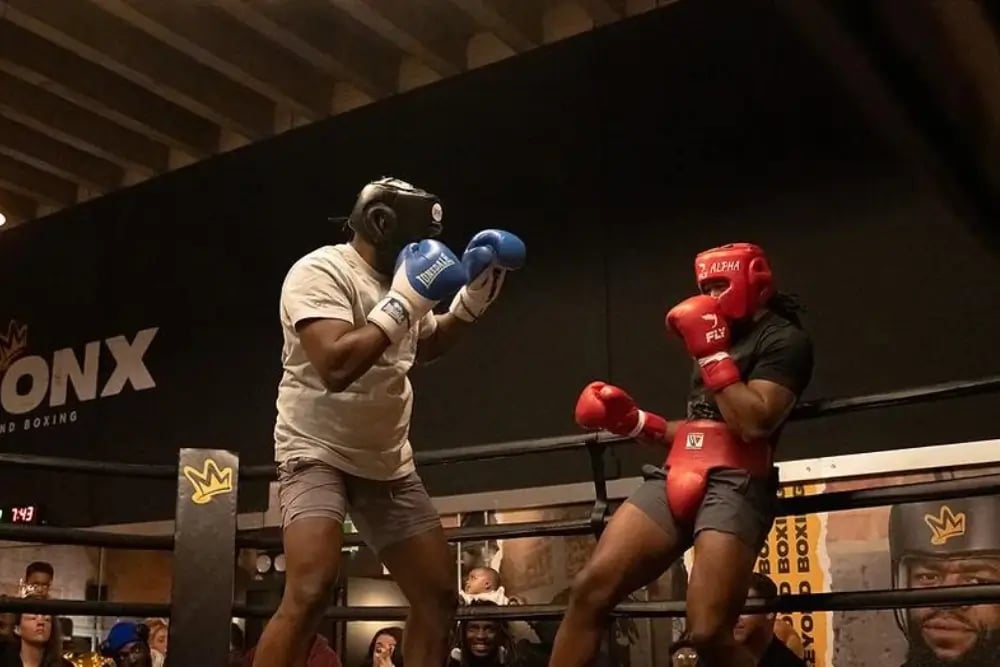 two fighters in a boxing ring at Bronx Boxing Club in London one of LoudLocal's SEO and digital strategy clients, one has white top, shorts, blue boxing gloves and black protective headgear, the second is leaning back on the ropes with black top, shorts, red boxing gloves and red headgear