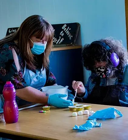 The Action Group is one of LoudLocal's clients. This image shows two people baking and spooning cake mix into cupcake cases, the are both wearing aprons and one has a pair of headphones on, they are doing this at one of the Action Group's support centres.