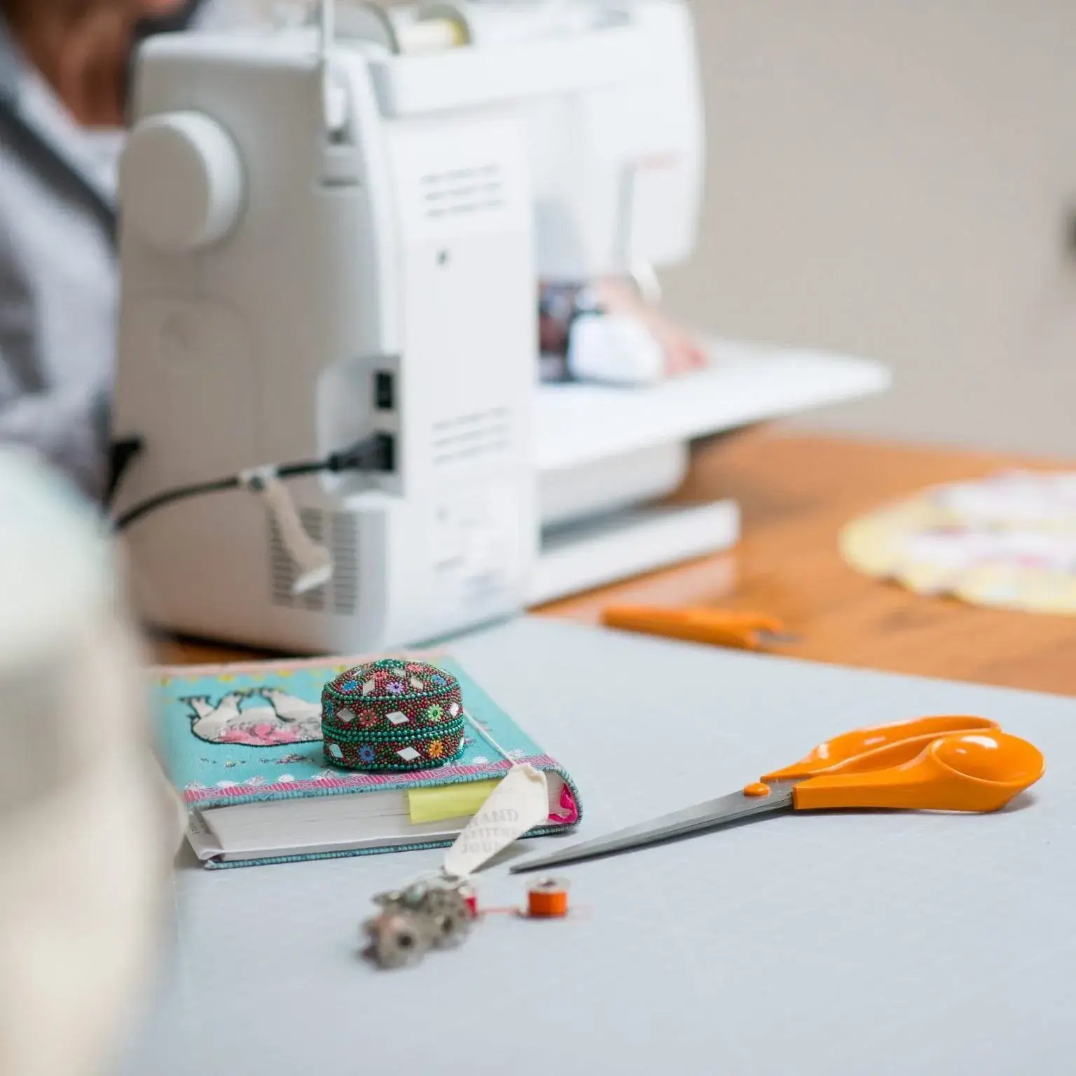 This image shows a sewing machine along with scissors with an orange handle and a blue notebook on a wooden table.