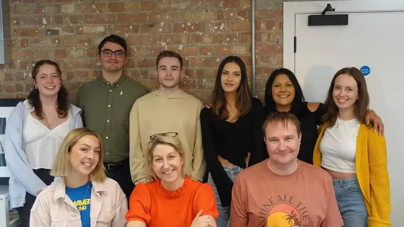 Nine LoudLocal colleagues posing together in an office beside an exposed brick wall.