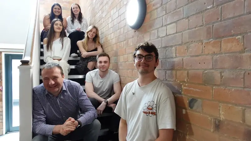 Seven LoudLocal colleagues sitting on the staircase at Spencer Yard in Leamington Spa, beside an exposed brick wall.