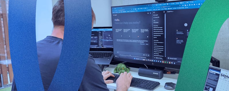 Leon our digital marketing director working at a PC using both Gemini and ChatGPT for AI Content generation as part of his SEO strategy. Leon is wearing a dark blue top, he has numerous LoudLocal branded merchandise on his desk. His desk is white with a black trim.