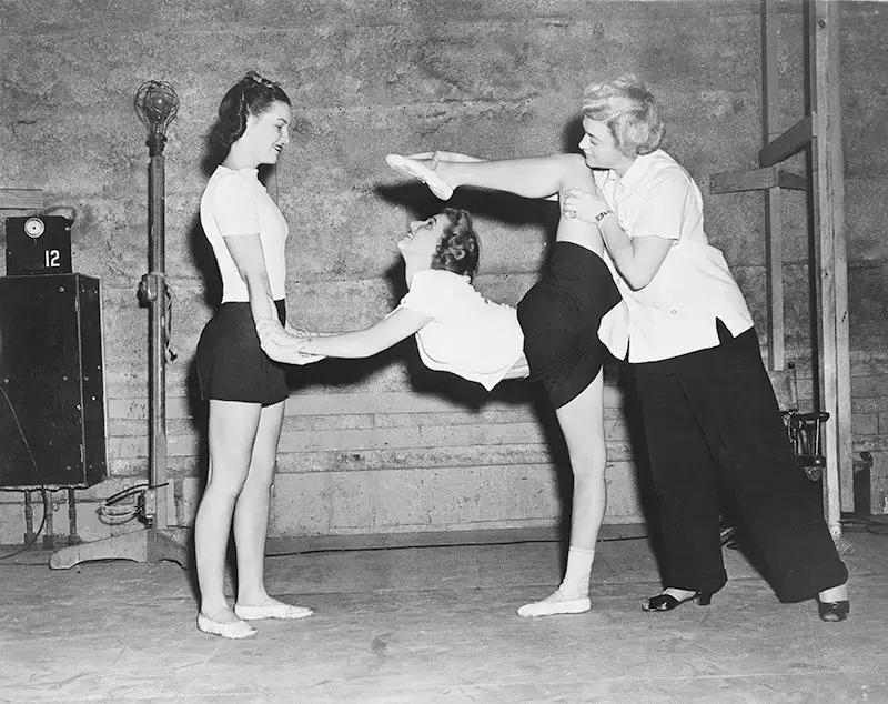 An image of 3 women, two are helping the one in the middle lift her leg over to touch her head
