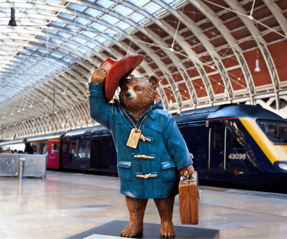 A model of the world famous Paddington bear at London station, he is wearing a light blue coat and is tipping his red hat as if to say hello.  He has a brown suitcase and wears a luggage tag round his neck. There is a dark blue train behind him.  