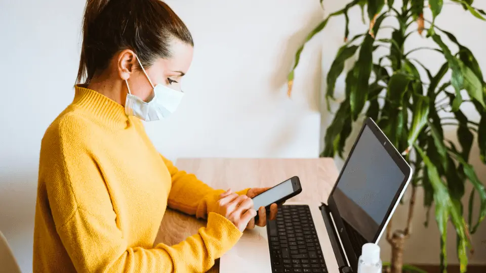 Woman in a yellow jumper using her phone whilst on a laptop and wearing a protective face mask