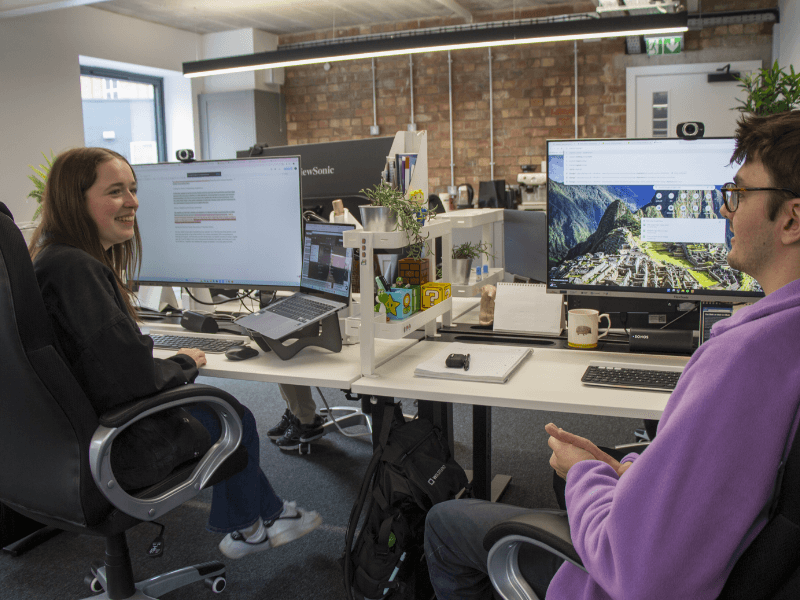 Toby and Mia sat at their desks discussing SEO strategy.  There is a brick wall in the background and the desks are whit e with a black trim.  Toby is wearing a purple hoodie and Mia is wearing a dark top and white trainers.  Both are quite young, Mia looks around 19 and Toby about 27.  There are windows in the background. 